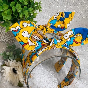 May include: A clear plastic headband with a fabric bow featuring a repeating pattern of yellow cartoon characters with blue and white accents. The headband is a fun accessory.