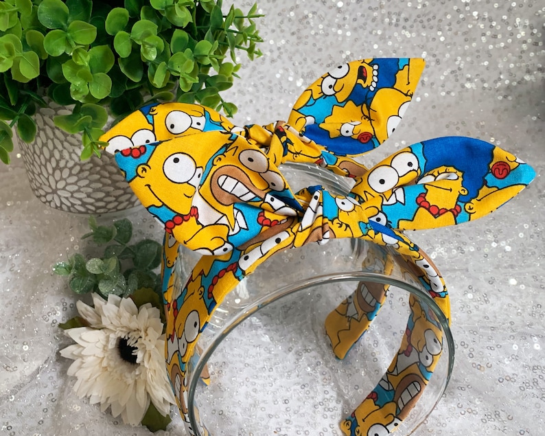 May include: A clear plastic headband with a fabric bow featuring a repeating pattern of yellow cartoon characters with blue and white accents. The headband is a fun accessory.