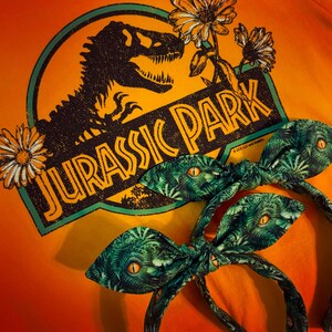 Pu&ograve; includere: Un cerchietto verde e nero con una stampa a occhio di dinosauro. Il cerchietto &egrave; legato a fiocco e si trova davanti a una maglietta gialla con il logo di Jurassic Park.