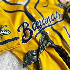 May include: Yellow baseball jersey with navy blue accents and the word "Bananas" in large script. A matching headband with a banana pattern is also visible. The jersey has the number 1 on it.