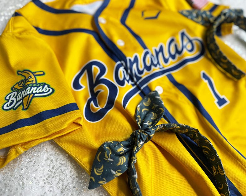 May include: Yellow baseball jersey with navy blue accents and the word "Bananas" in large script. A matching headband with a banana pattern is also visible. The jersey has the number 1 on it.