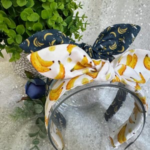 May include: A clear plastic headband with a white fabric bow featuring yellow bananas. A second headband with a dark blue bow with yellow banana prints is also visible. Green foliage and a blue flower are in the background.