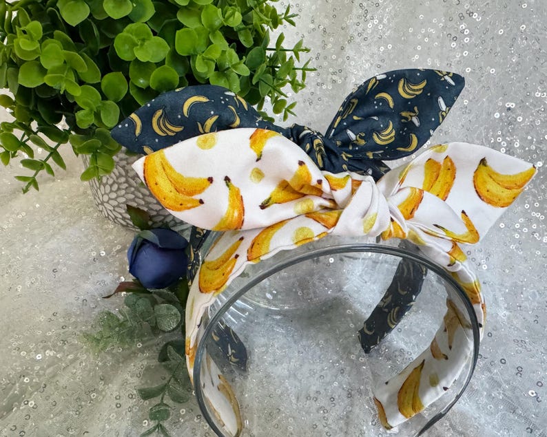 May include: A clear plastic headband with a white fabric bow featuring yellow bananas. A second headband with a dark blue bow with yellow banana prints is also visible. Green foliage and a blue flower are in the background.