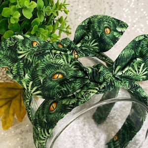 Pu&ograve; includere: Fascia per capelli in tessuto verde e giallo con un nodo al centro. Il tessuto ha un motivo di foglie verdi e occhi gialli.