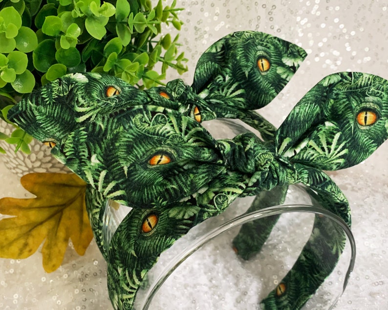 Pu&ograve; includere: Fascia per capelli in tessuto verde e giallo con un nodo al centro. Il tessuto ha un motivo di foglie verdi e occhi gialli.