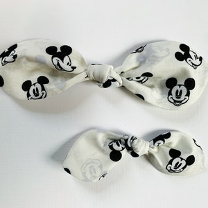 May include: Two white fabric hair bows with a black and white Mickey Mouse print. The larger bow is tied in a knot, while the smaller bow is tied in a simple bow.