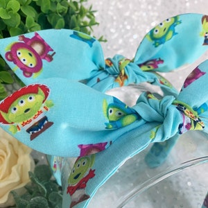 May include: A light blue headband with a repeating pattern of green alien characters from the Toy Story franchise. The headband is tied in a bow at the top.