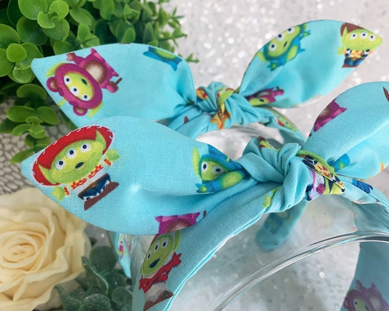 May include: A light blue headband with a repeating pattern of green alien characters from the Toy Story franchise. The headband is tied in a bow at the top.