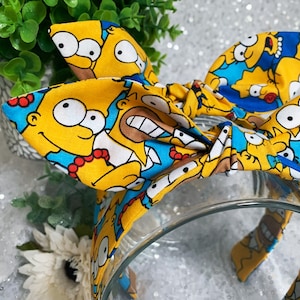May include: A yellow headband featuring a repeating pattern of Homer Simpson's face. The headband has a knotted bow design and is set against a blurred background with greenery and a clear glass bowl.