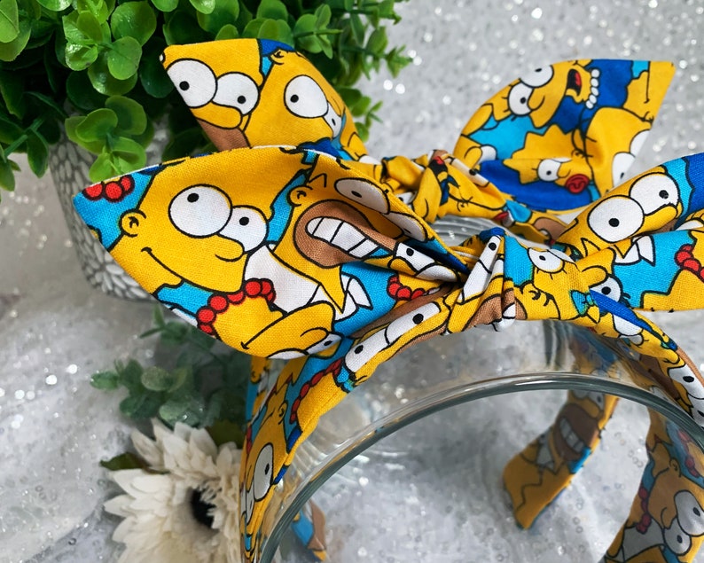 May include: A yellow headband featuring a repeating pattern of Homer Simpson's face. The headband has a knotted bow design and is set against a blurred background with greenery and a clear glass bowl.