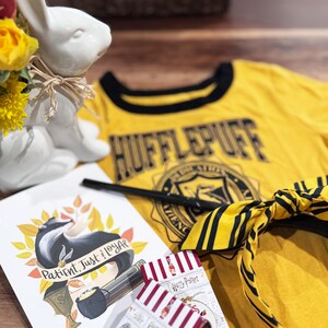 May include: A yellow Hufflepuff t-shirt with black trim, a wand, and a yellow and black striped scarf. A white ceramic bunny vase holds yellow flowers. A card with a badger illustration and the words "Patient, Just, Loyal" is also present.