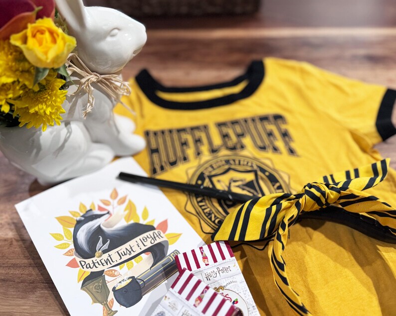 May include: A yellow Hufflepuff t-shirt with black trim, a wand, and a yellow and black striped scarf. A white ceramic bunny vase holds yellow flowers. A card with a badger illustration and the words "Patient, Just, Loyal" is also present.