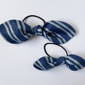 May include: Two blue and white striped fabric hair scrunchies. The larger scrunchie is tied in a bow and the smaller scrunchie is tied in a bow.