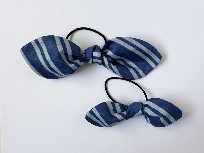 May include: Two blue and white striped fabric hair scrunchies. The larger scrunchie is tied in a bow and the smaller scrunchie is tied in a bow.
