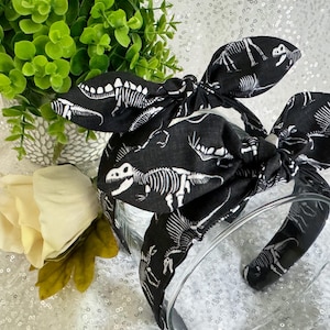 May include: Black headband with a white dinosaur skeleton print. The headband is tied in a bow at the top.