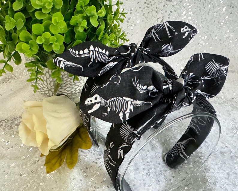 May include: Black headband with a white dinosaur skeleton print. The headband is tied in a bow at the top.