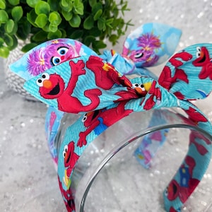 May include: A fabric headband with a bow, patterned with Elmo and other Sesame Street characters on a light blue background. The headband has a matching pattern.