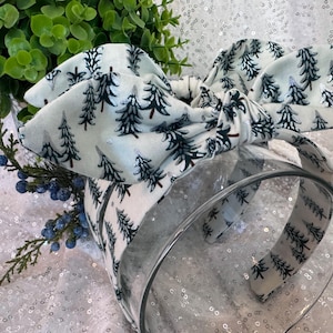 May include: A fabric headband with a bow, patterned with dark green fir trees on a white background. The headband is displayed with greenery and a clear glass container, creating a festive look.