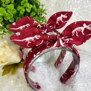 May include: Red headband with a white dinosaur skeleton print. The headband is tied in a bow at the top.