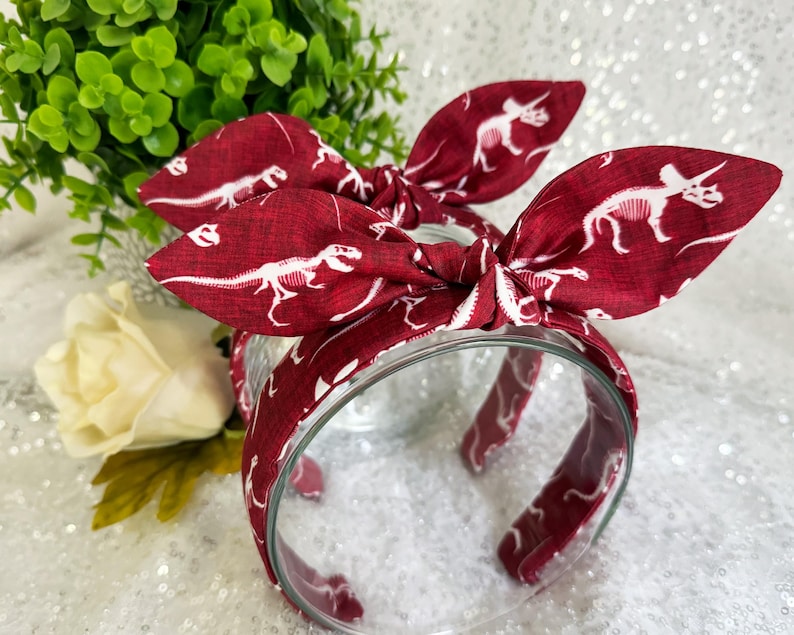 May include: Red headband with a white dinosaur skeleton print. The headband is tied in a bow at the top.