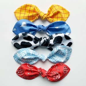 May include: Five fabric hair bows in different patterns. The top bow is yellow and red checked, the second is blue with white clouds, the third is black and white cow print, the fourth is blue with a white geometric pattern, and the bottom bow is red with a white bandana pattern.