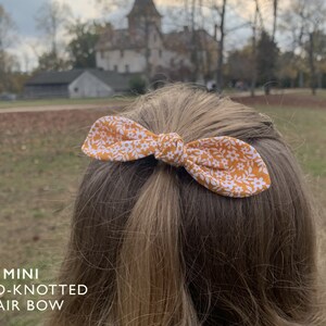 K&ouml;nnte beinhalten: Ein kleiner, handgekn&uuml;pfter Haarbogen mit einem gelben und wei&szlig;en Blumenmuster. Die Schleife ist in einem einfachen Knoten gebunden und wird im Haar einer Person getragen.