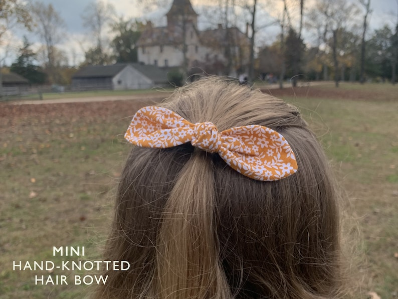 K&ouml;nnte beinhalten: Ein kleiner, handgekn&uuml;pfter Haarbogen mit einem gelben und wei&szlig;en Blumenmuster. Die Schleife ist in einem einfachen Knoten gebunden und wird im Haar einer Person getragen.