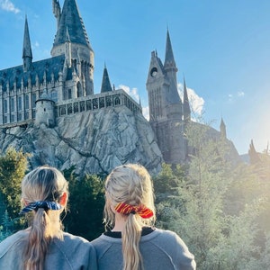 May include: Two people stand in front of a large stone castle with tall towers. The castle is gray and has many windows. The people are wearing casual clothing and have their hair pulled back. The sky is blue and there are trees in the background.