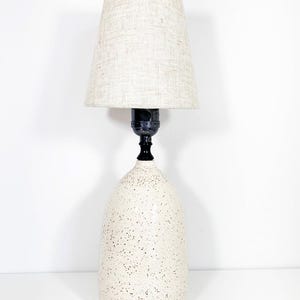 May include: A cream-colored table lamp with a speckled base and a beige linen lampshade. The lamp has a black neck and a bulb socket. The lamp is a decorative lighting fixture.