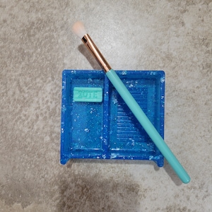 Könnte beinhalten: Ein blaues Glitzertablett mit einem kleinen grünen Seifenstück, auf dem "VOTE" steht, und einem hellblauen Make-up-Pinsel.