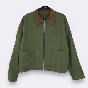 May include: Olive green jacket with a brown collar and a silver zipper. The jacket has two front pockets and is hanging on a black hanger. The jacket is a casual style.