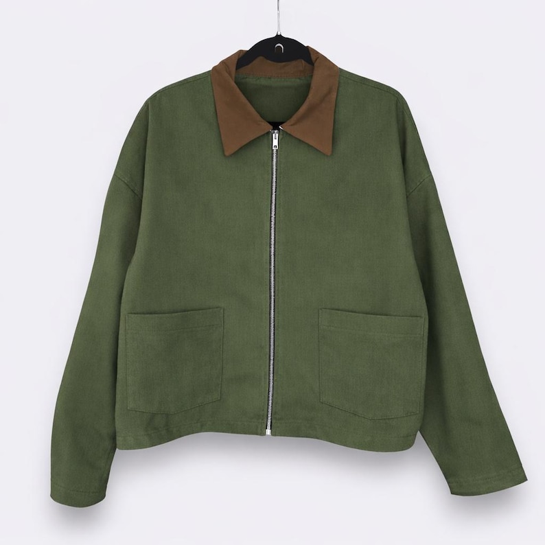 May include: Olive green jacket with a brown collar and a silver zipper. The jacket has two front pockets and is hanging on a black hanger. The jacket is a casual style.