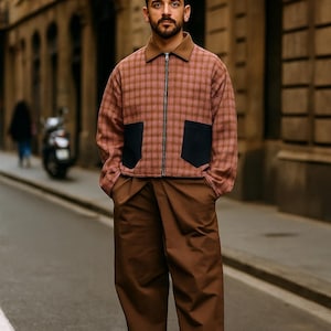 May include: A person wearing a brown and pink plaid jacket with black pockets, a brown collar, and brown pants. The jacket has a zipper closure. The person is standing on a city street.