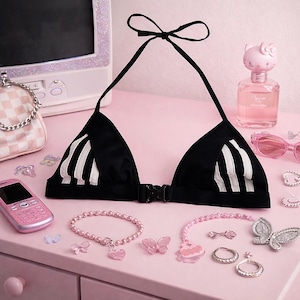 Puede incluir: Top de bikini negro con rayas blancas, exhibido con accesorios rosas que incluyen una pulsera, pendientes, gafas de sol y un teléfono de estilo retro. También se ven un frasco de perfume rosa y un pequeño bolso.