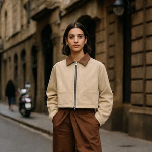 May include: A beige jacket with a brown collar and a zipper closure. The jacket has two front pockets and is paired with brown pants. The person in the image is standing on a city street.