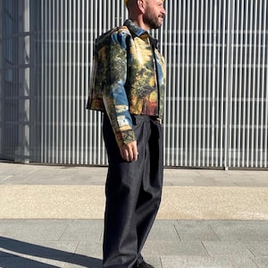 Puede incluir: Una persona con una chaqueta estampada con un paisaje, pantalones oscuros y un gorro amarillo. La chaqueta presenta un diseño escénico colorido con árboles y agua. El atuendo se completa con zapatos negros. El fondo es un edificio moderno.