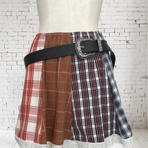 Puede incluir: Minifalda patchwork con una variedad de patrones de cuadros en rojo, marrón y azul. La falda está adornada con un cinturón negro con una hebilla plateada decorativa. El dobladillo está rematado con un ribete de encaje blanco.