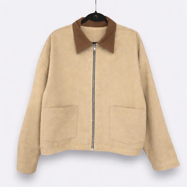 May include: A tan jacket with a brown collar and a full-length zipper. The jacket has two front pockets and is hanging on a black hanger. The jacket is made of a soft material.