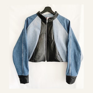 Könnte beinhalten: Eine verkürzte Jacke aus Denim und Leder mit Reißverschluss. Die Jacke hat ein schwarzes Lederpanel in der Mitte und Denim-Panels an den Seiten und Ärmeln. Die Ärmel haben schwarze Ledermanschetten.