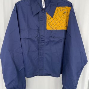 Peut inclure: Veste bleu marine avec deux poches avant et un écusson jaune représentant un visage dans un motif quadrillé. La veste a des manches longues avec des poignets élastiques et un col. L'étiquette indique "FRANKS".