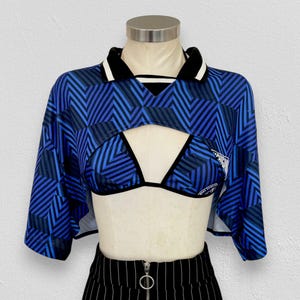 Könnte beinhalten: Ein blau-schwarzes bauchfreies Oberteil mit geometrischem Zickzackmuster, schwarzem Kragen mit weißem Rand und einem passenden Bikini-Oberteil darunter. Das Kleidungsstück wird auf einer Schaufensterpuppe mit schwarzer Nadelstreifenhose präsentiert.