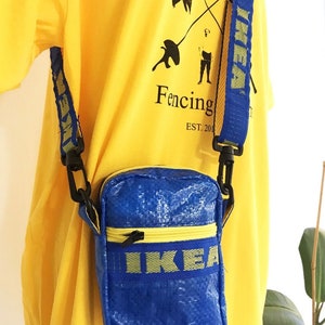 May include: A blue crossbody bag with a yellow zipper and strap, featuring the IKEA logo. The bag is worn over a yellow t-shirt with a black graphic and the text "Fencing EST. 2018".