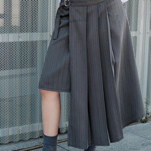 Puede incluir: Una falda cruzada con estampado de espiga en gris oscuro y blanco, con un panel lateral plisado, cierre de hebilla ajustable en la cintura y dobladillo asimétrico. La falda se combina con una camisa blanca, calcetines grises y zapatos negros.
