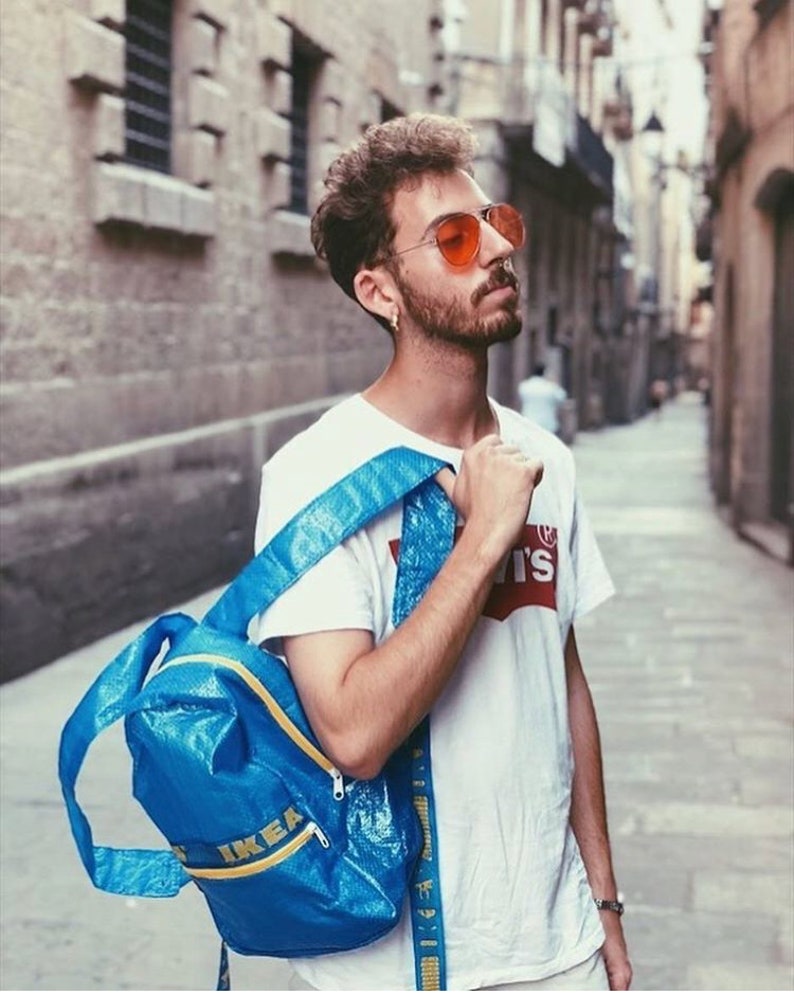 K&ouml;nnte beinhalten: Eine Person tr&auml;gt ein wei&szlig;es T-Shirt mit dem Aufdruck "Levi's", eine orangefarbene Sonnenbrille und einen blauen IKEA-Rucksack.