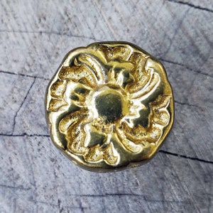 May include: A gold-toned metal knob with a floral design. The knob has a raised, ornate pattern and a smooth, shiny finish.