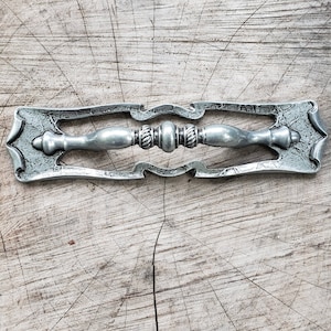 Pode incluir: Uma puxador de gaveta de prata com um design ornamentado. O puxador tem uma pega central com um botão decorativo em cada extremidade. O puxador está colocado sobre um fundo de madeira.