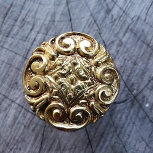 May include: A gold-toned metal knob with a decorative design of swirling patterns. The knob has a raised, diamond-shaped center with intricate details.