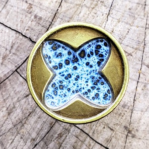 Op de afbeelding: Een ronde, messingkleurige knoop met een blauw en wit, ingewikkeld, fractal-achtig ontwerp in het midden.