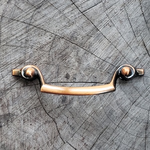 May include: A copper-toned drawer pull with a curved design and ornate details. The pull is mounted on a wooden surface.