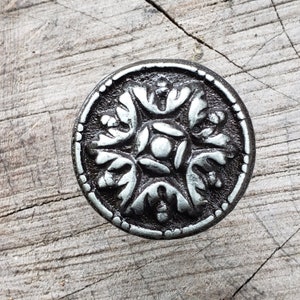 May include: A silver metal button with a decorative floral design. The button has a raised, circular border and a central design of four stylized flowers.